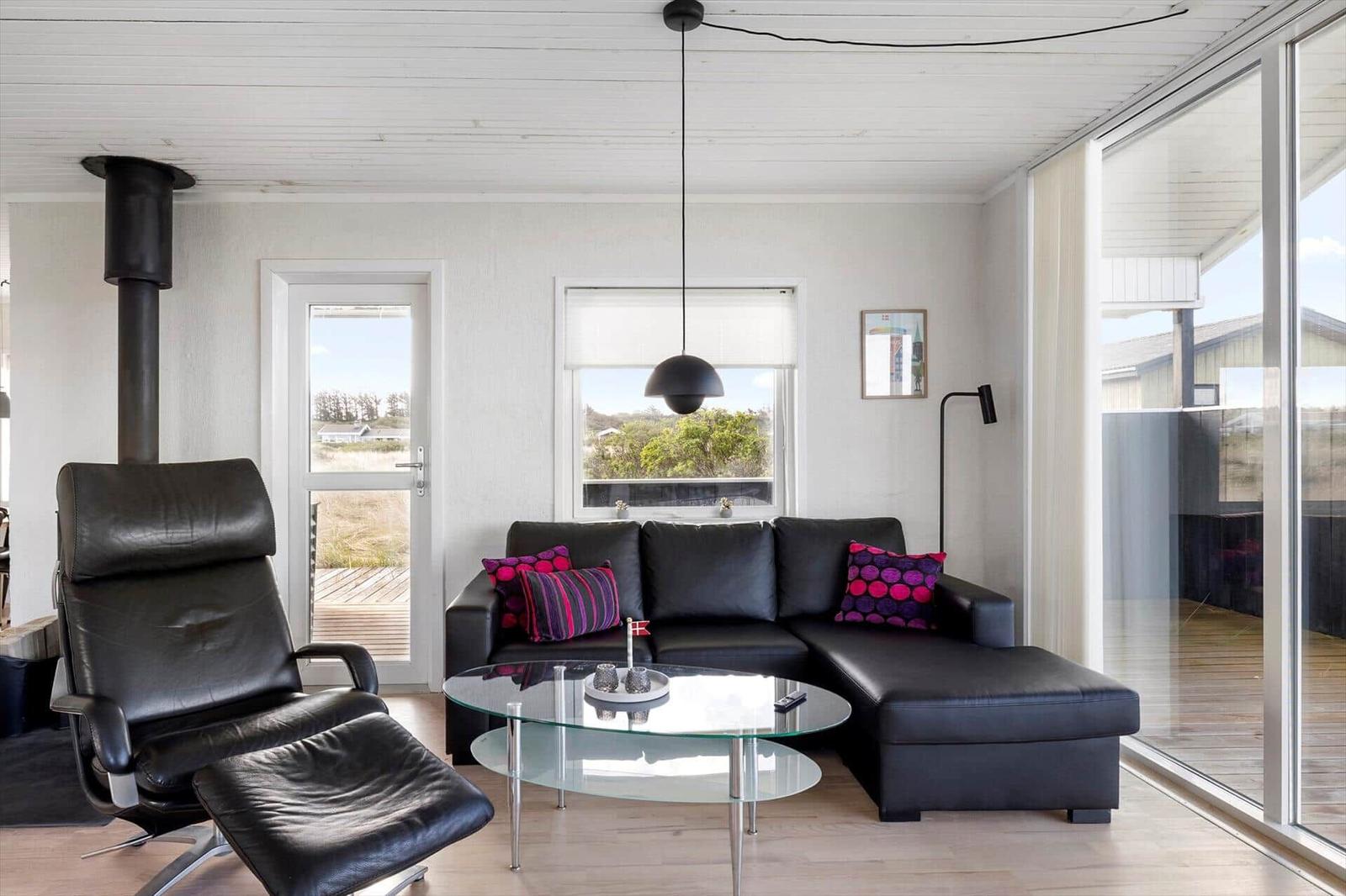 Living room with black leather sofa, glass table, and seating. Large windows and door with view of outdoor area.