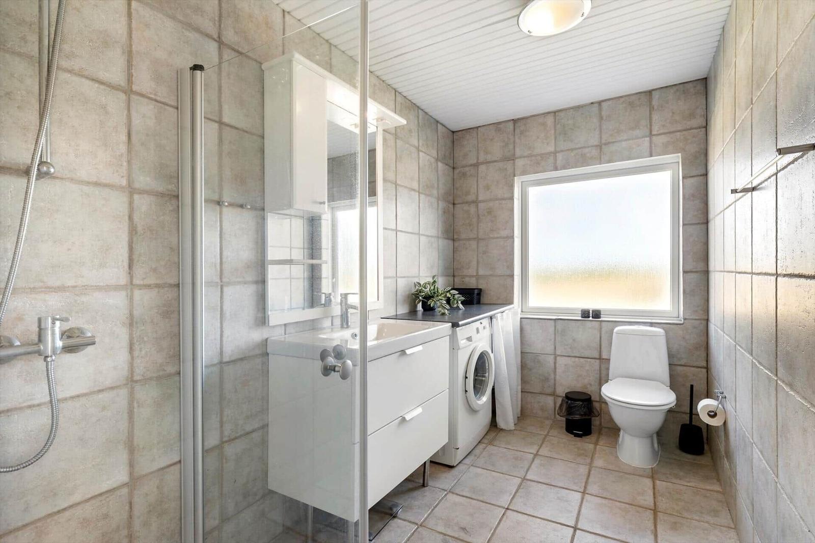 Bathroom with shower, washing machine, and toilet. Tiled walls and window.
