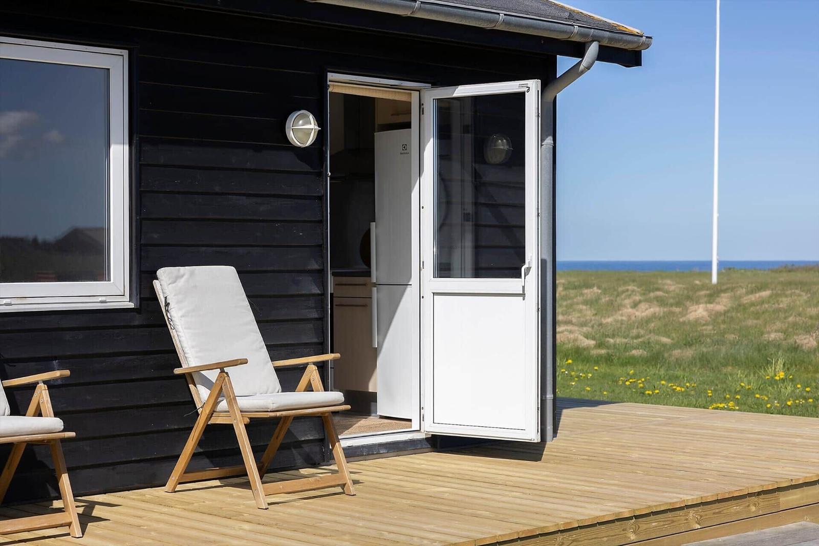 Schwarze Hütte mit Terrasse, weiße Tür und Stühle, Blick auf Grasland und Meer.