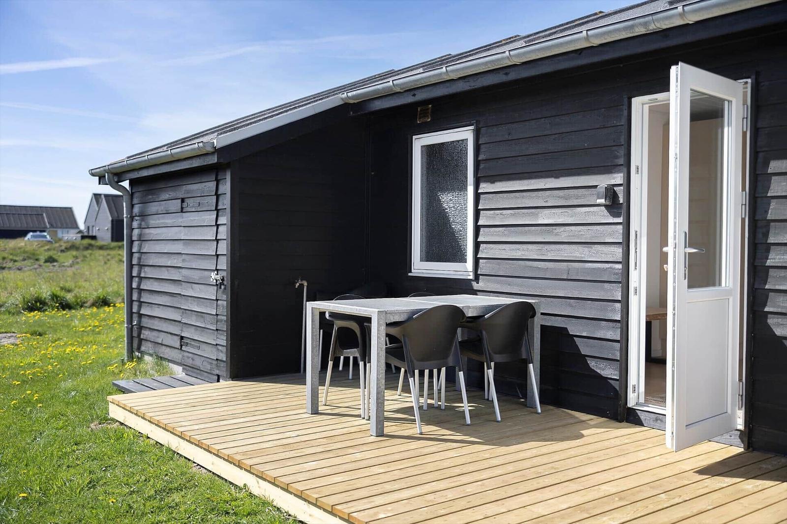 Schwarzes Holzhaus mit Terrasse, Tisch und Stühlen. Tür offen. Grasland im Hintergrund.