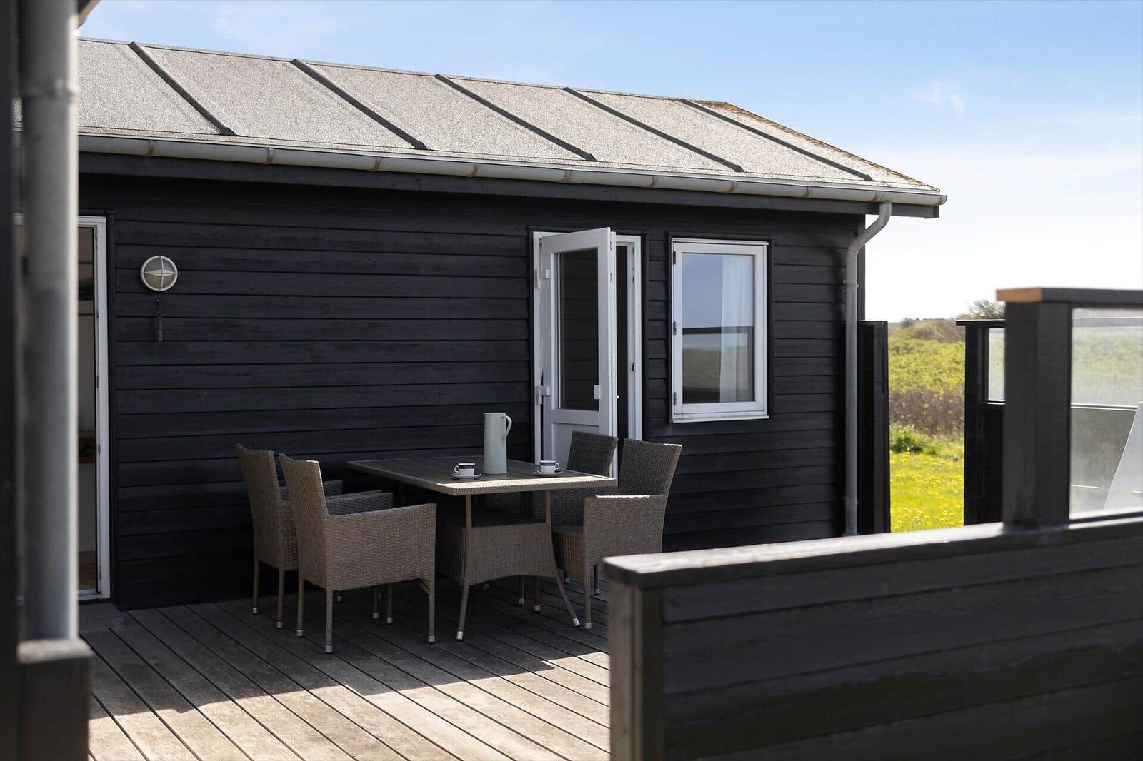 Terrasse mit Tisch und Stühlen vor einem dunklen Holzhaus mit offener Tür.