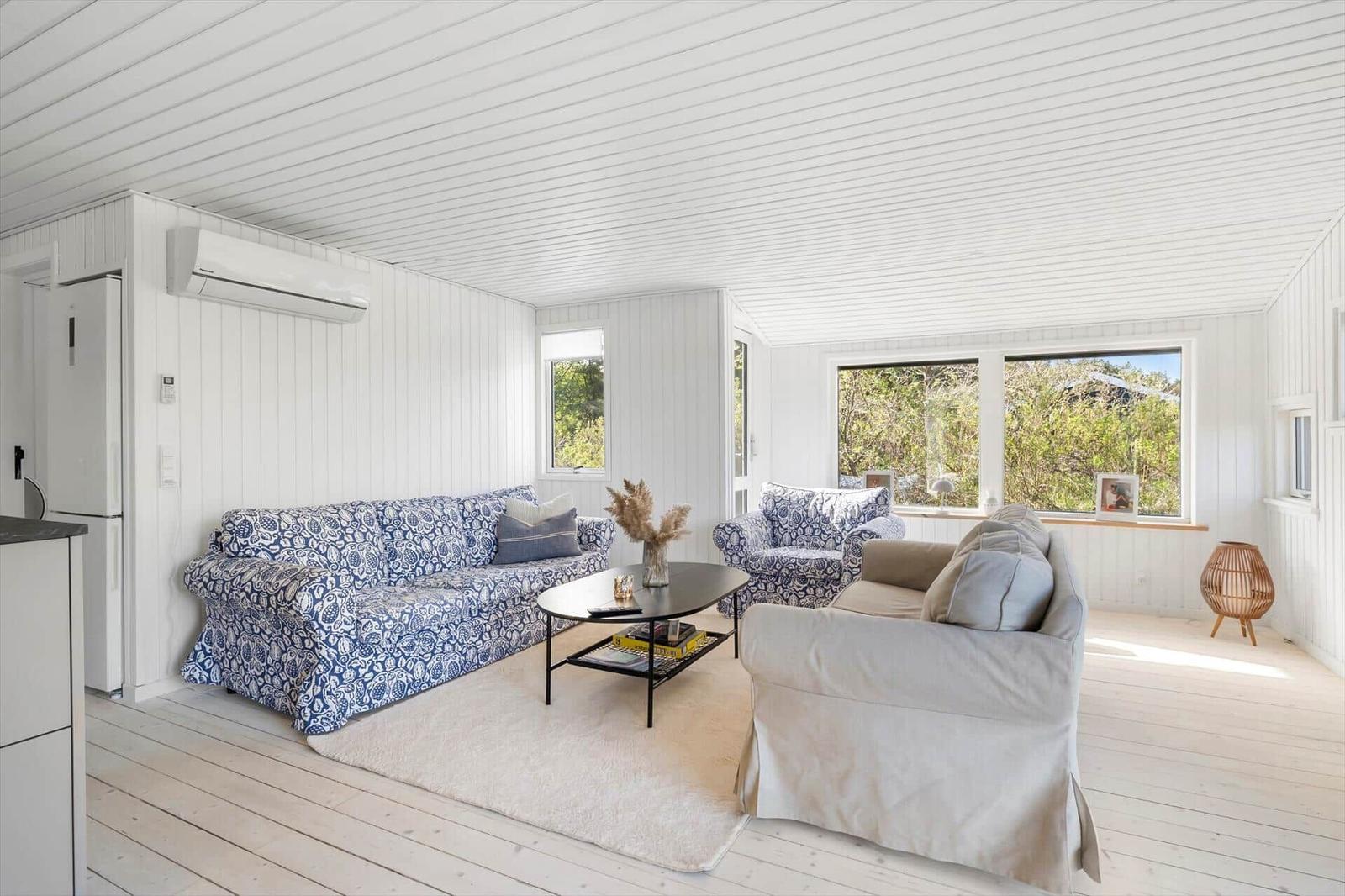 Living room with sofa, armchair, and table. Windows with view of greenery. White walls and ceiling.
