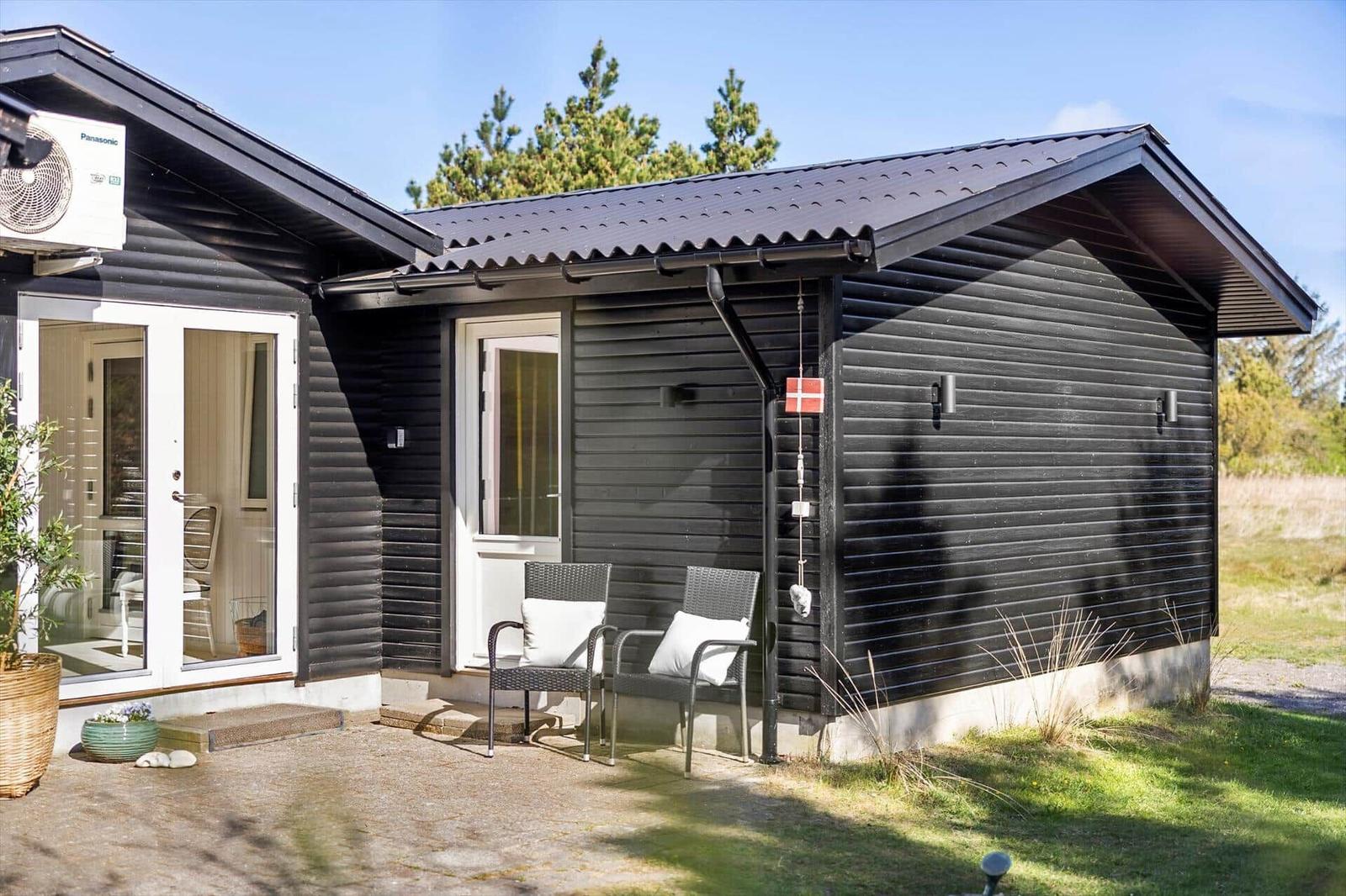 Black wooden cabin with terrace, two chairs, and garden doors. Danish flag hangs on the wall.