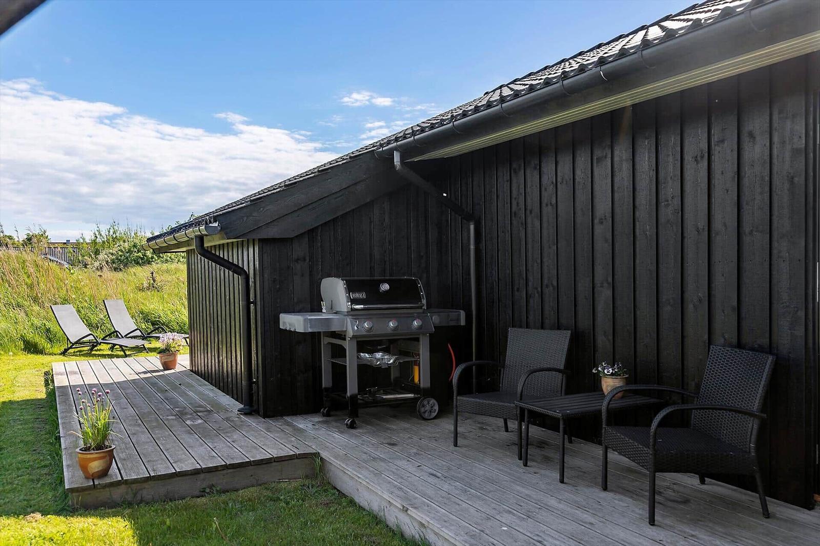 Deck with grill, two chairs and loungers beside a wooden house.