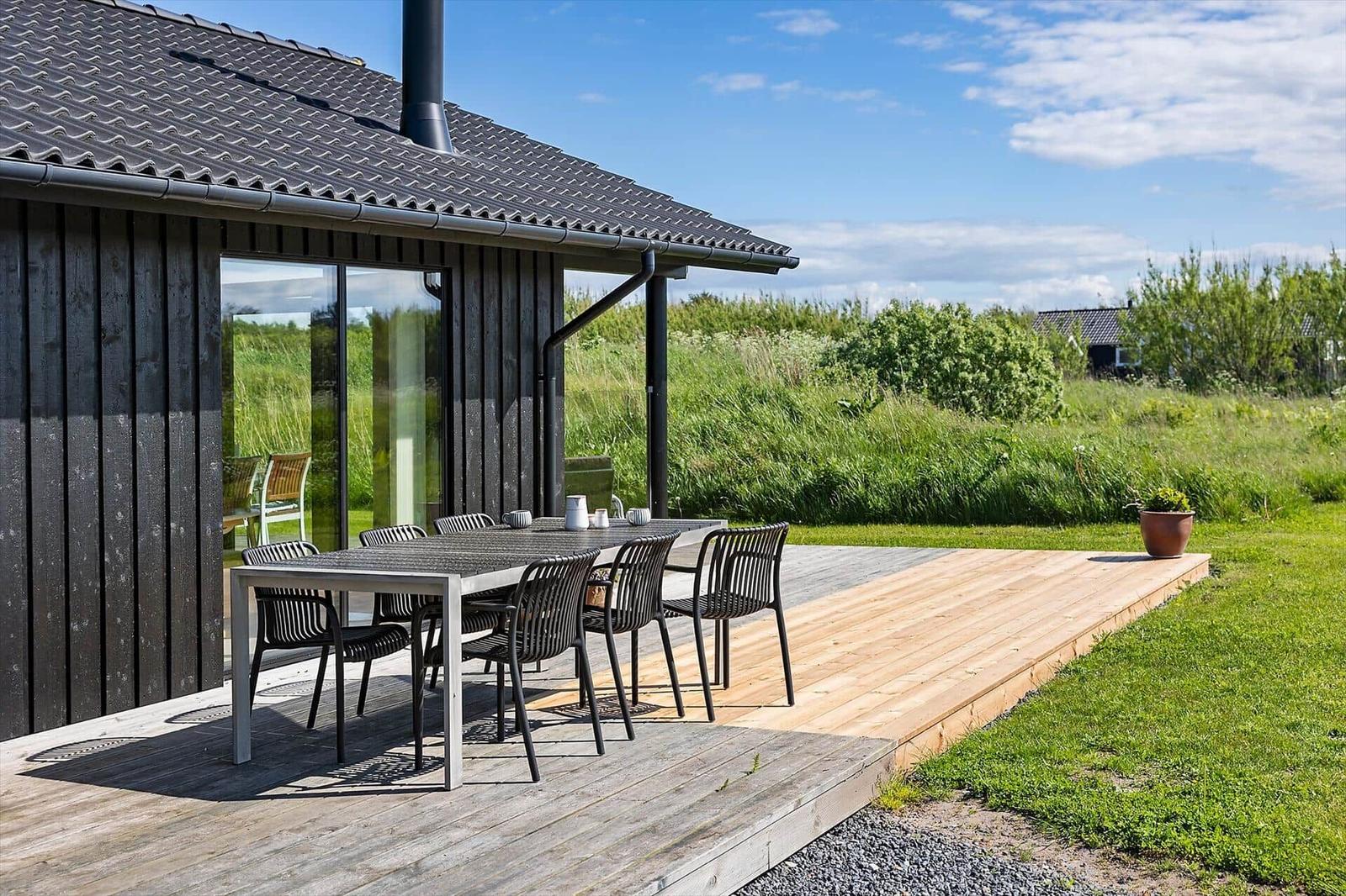 Deck with table and chairs in front of a house with glass facade and green background.