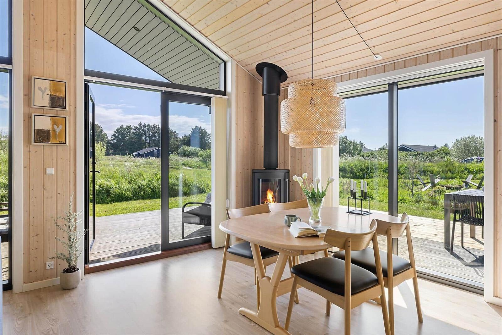 Dining room with wooden table, chairs, and fireplace. Large windows show garden and sky.