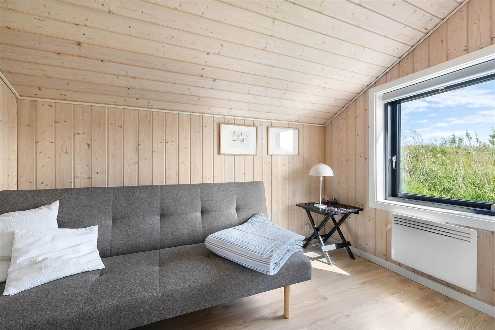 A room with wooden walls and ceiling, gray sofa, table, and window with grass landscape.
