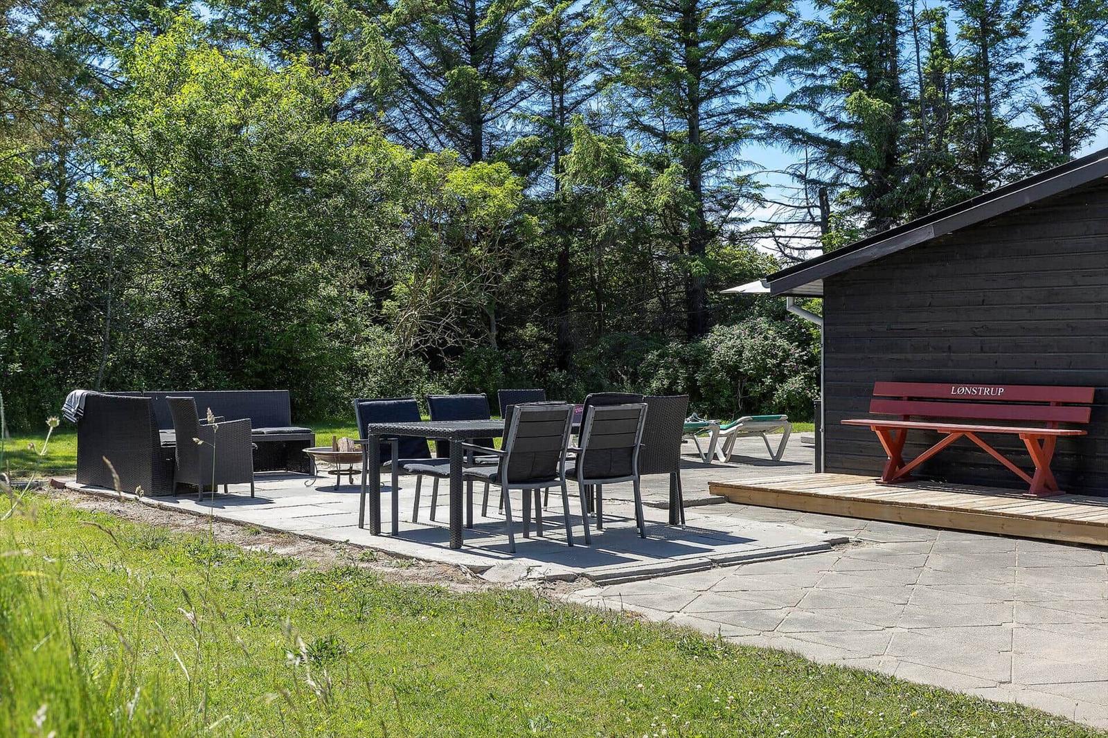 Terrasse mit Sitzgruppe, Tisch und Stühlen. Roter Bank neben Holzhaus. Grün hinter dem Haus.