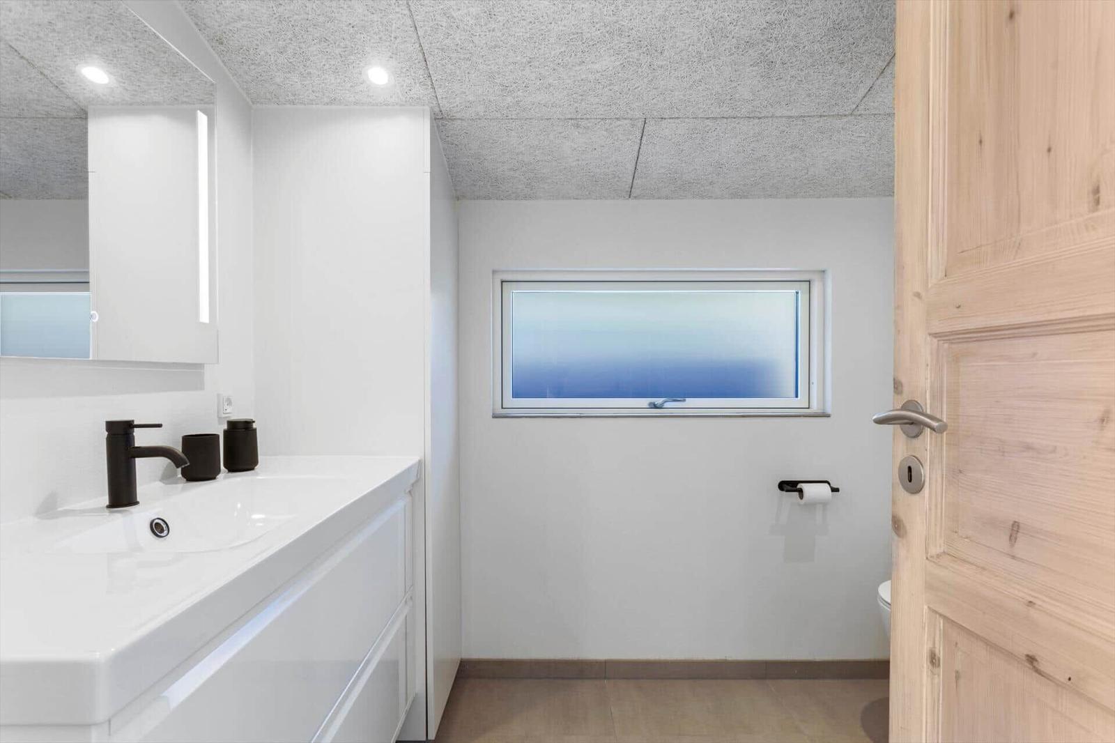 Badezimmer mit Waschtisch, Spiegel und Fenster. Holztür und Toilettenpapierhalter sichtbar.