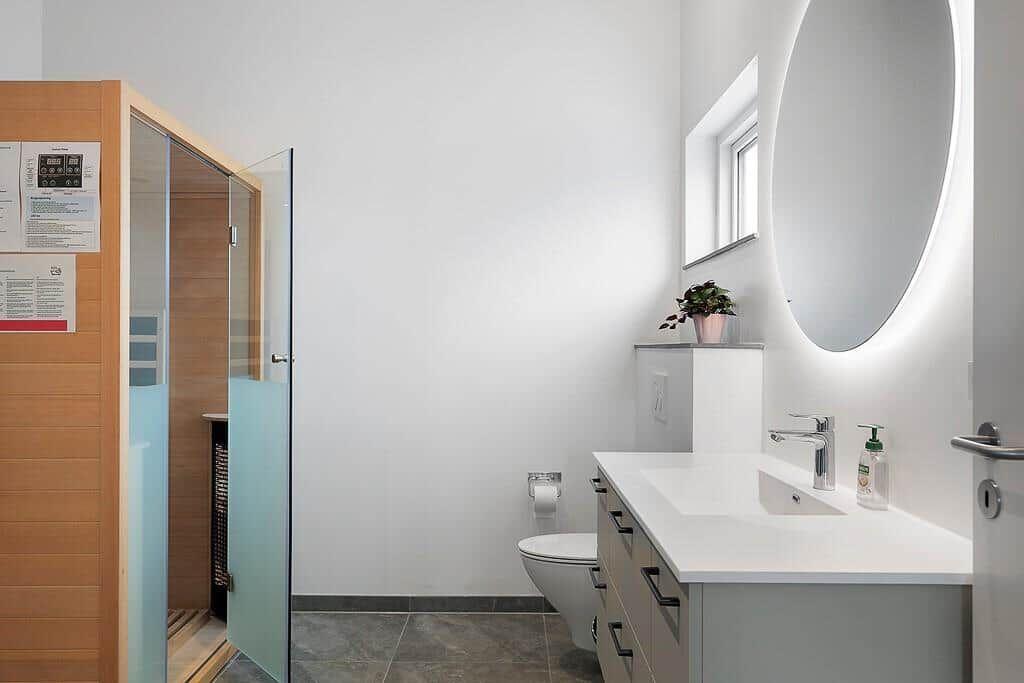 Badezimmer mit Sauna, Waschbecken und Toilette. Rundes Spiegel mit Beleuchtung.