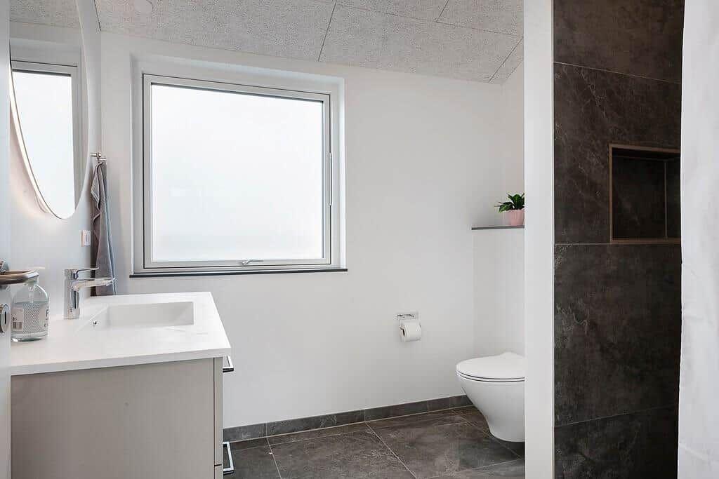 Badezimmer mit Waschbecken, Toilette und Fenster. Duschbereich mit dunkler Wandverkleidung.