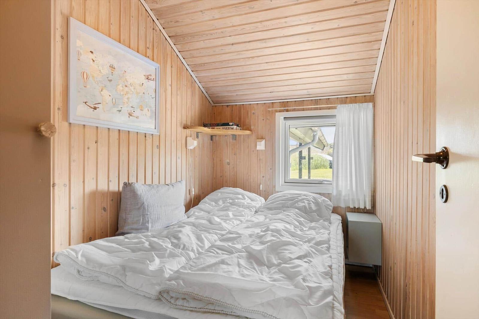 Schlafzimmer mit Holzwand, Bett und Fenster mit weißen Vorhängen.