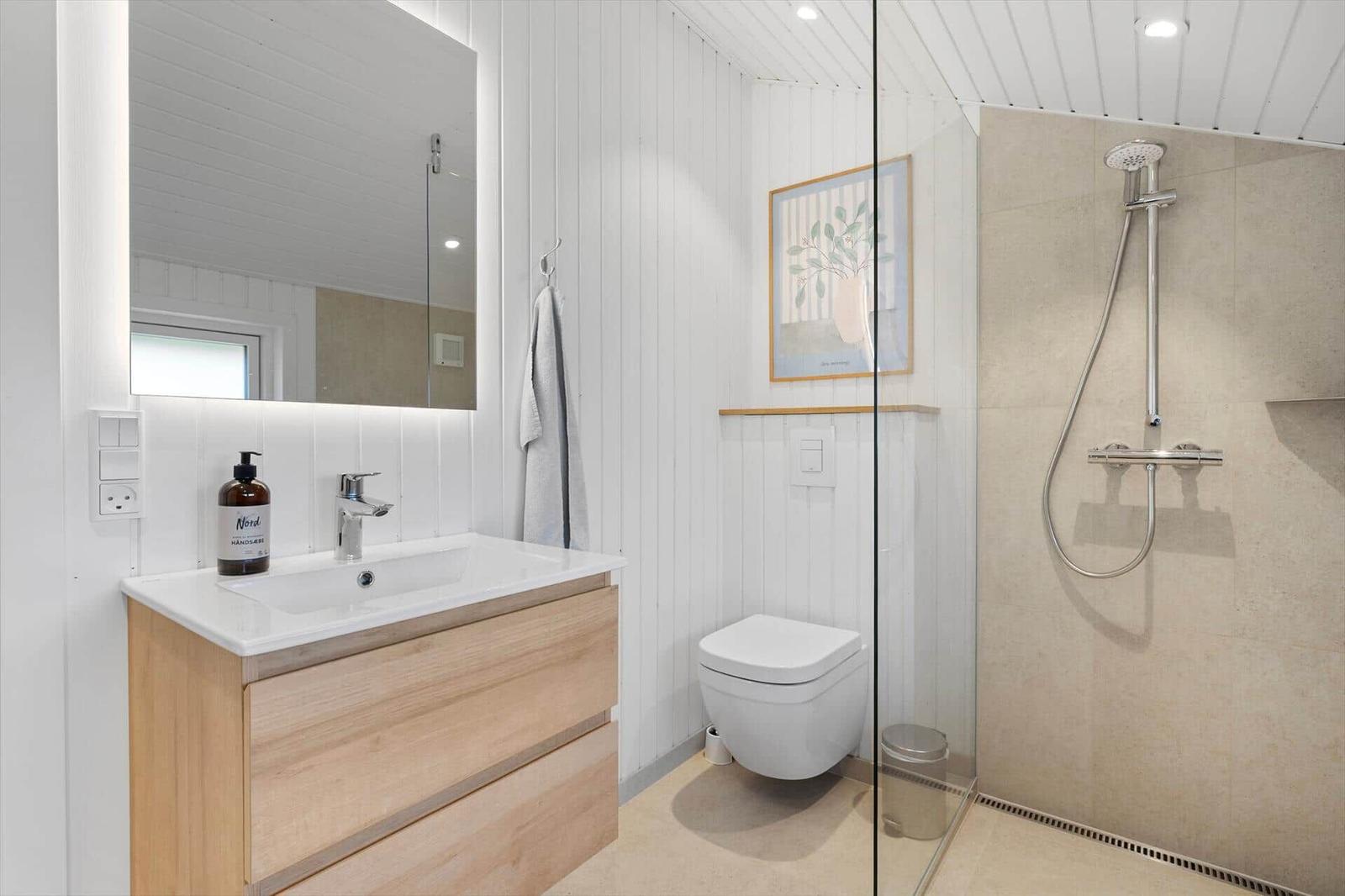Badezimmer mit Dusche, Waschbecken und Toilette. Holzunterschrank und Spiegel mit Beleuchtung.