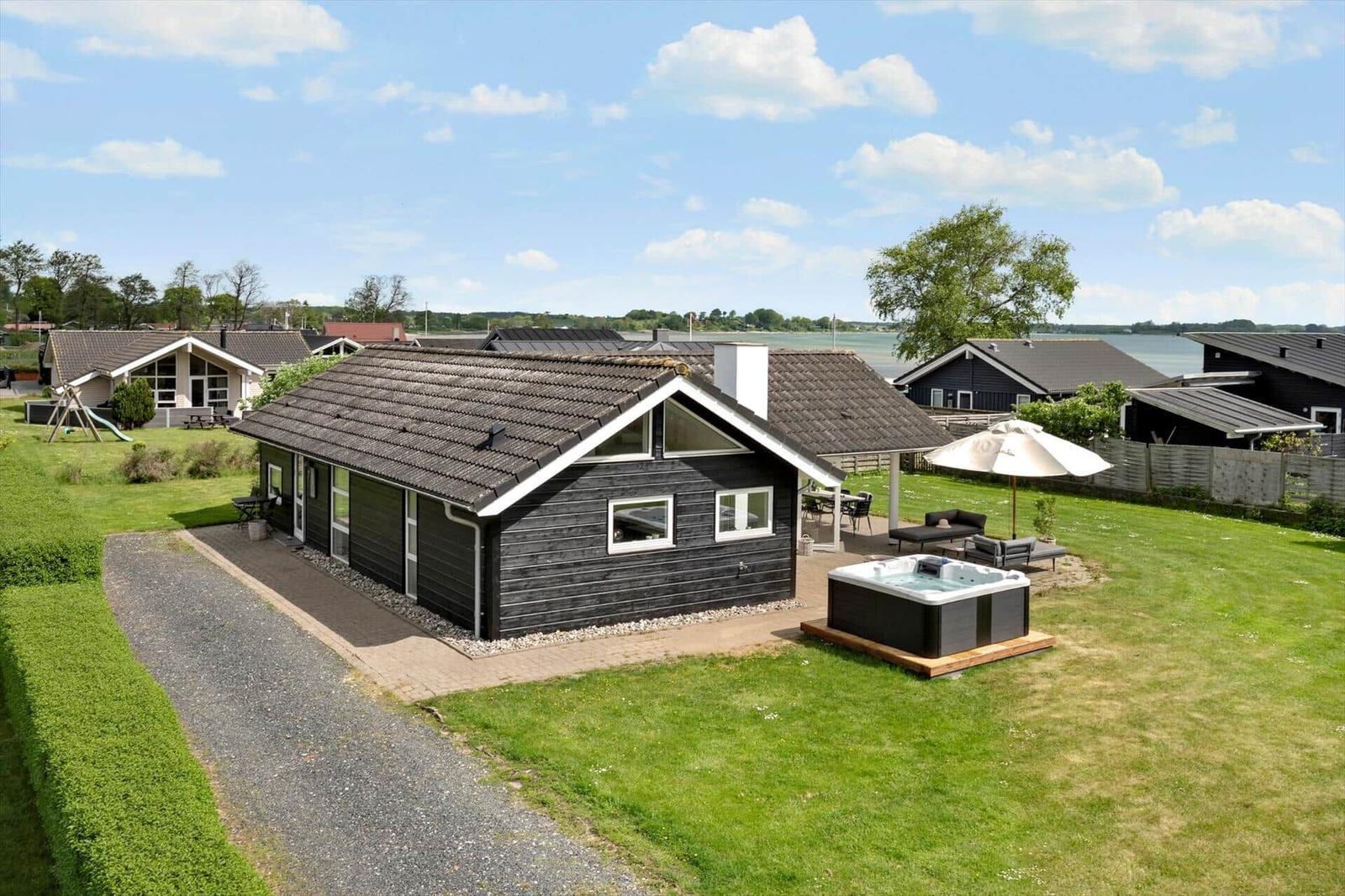 Black wooden house with hot tub and terrace. Lawn and water in the background.