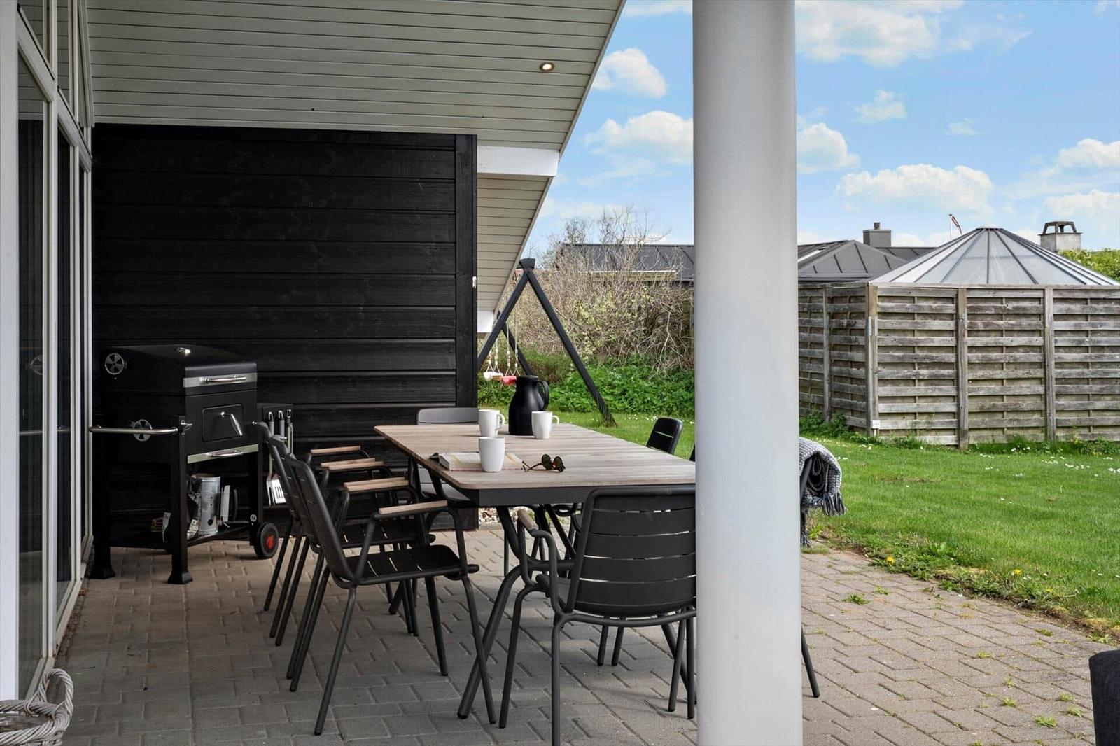 Patio with table, chairs, and grill under cover. Lawn and wooden fence in background.