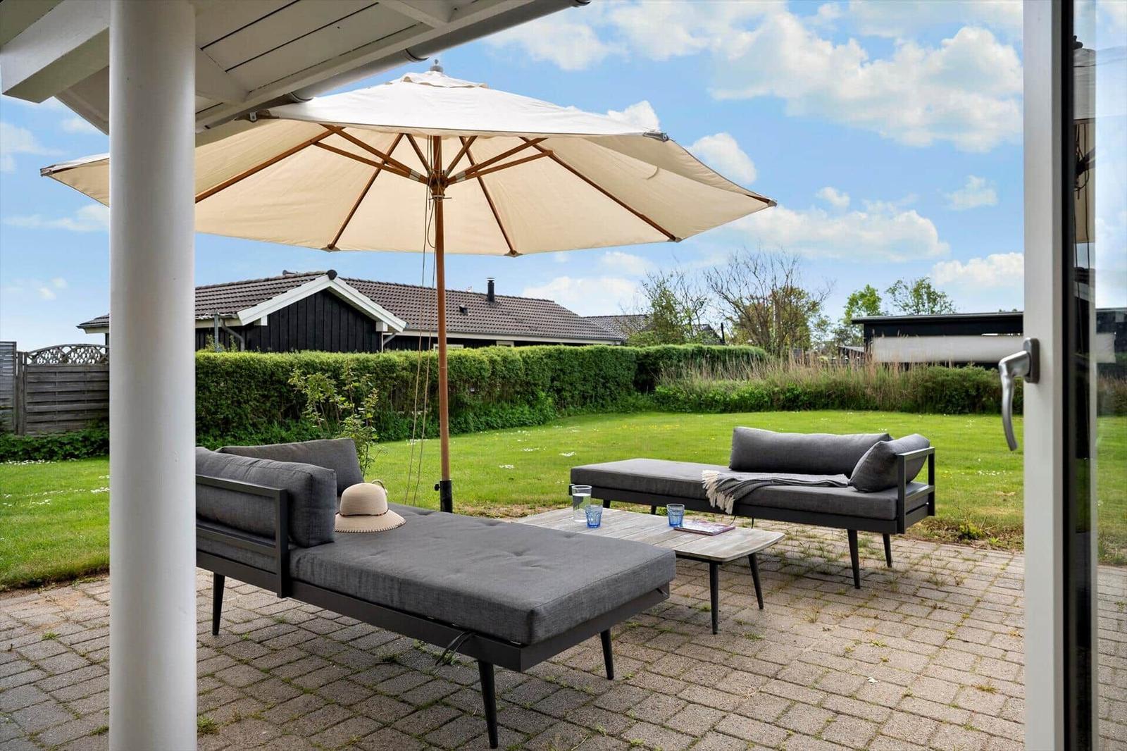Terrace with umbrella and two loungers on paved ground. Green lawn and hedge in the background.