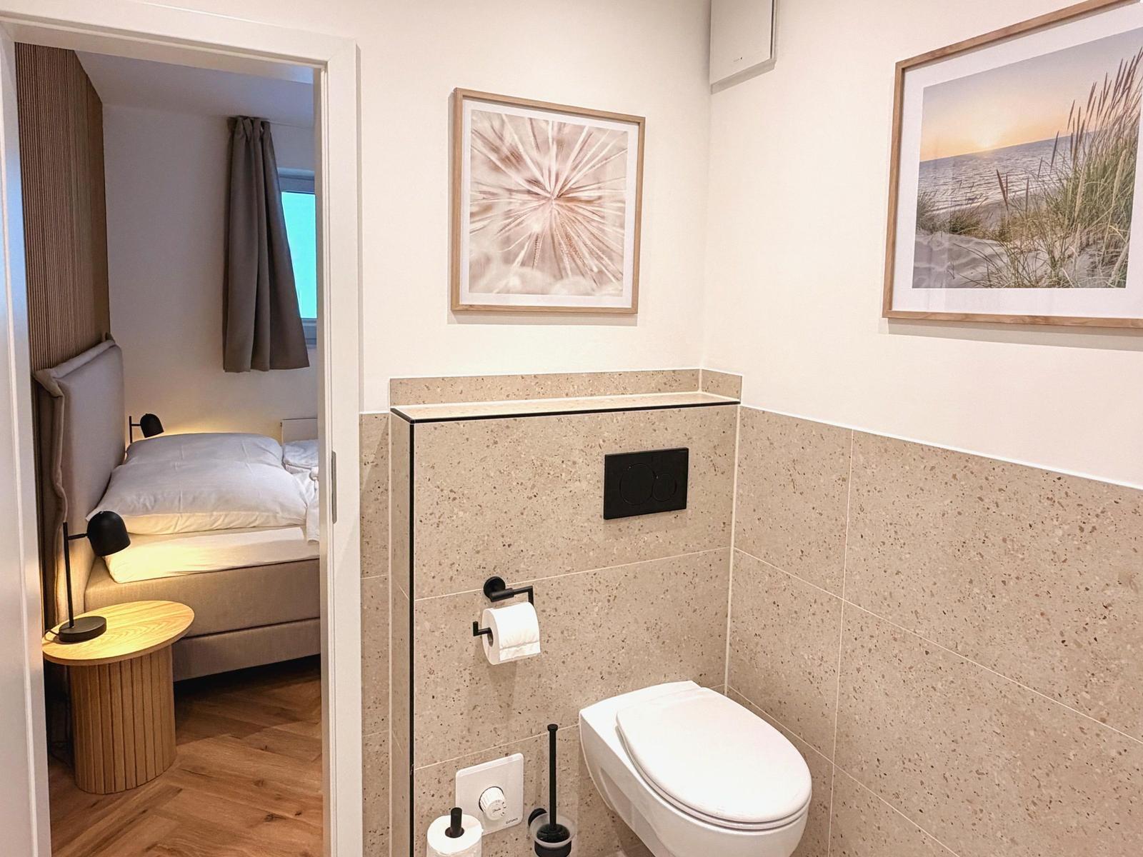 Badezimmer mit Toilette und Blick in Schlafzimmer mit Bett und Nachttisch.