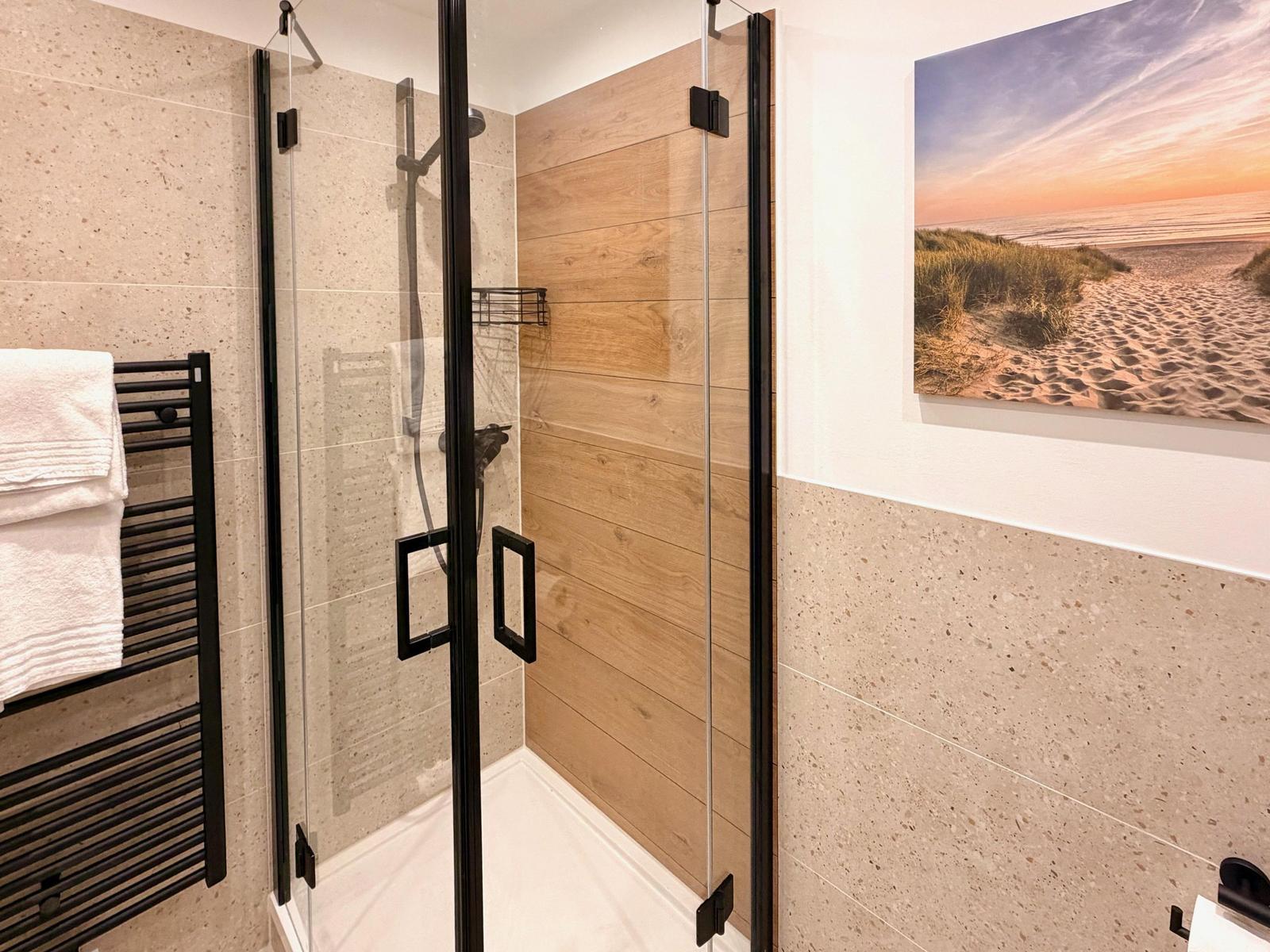 Duschkabine mit Glaswand und Holzwand, daneben Heizkörper und Strandbild an der Wand.
