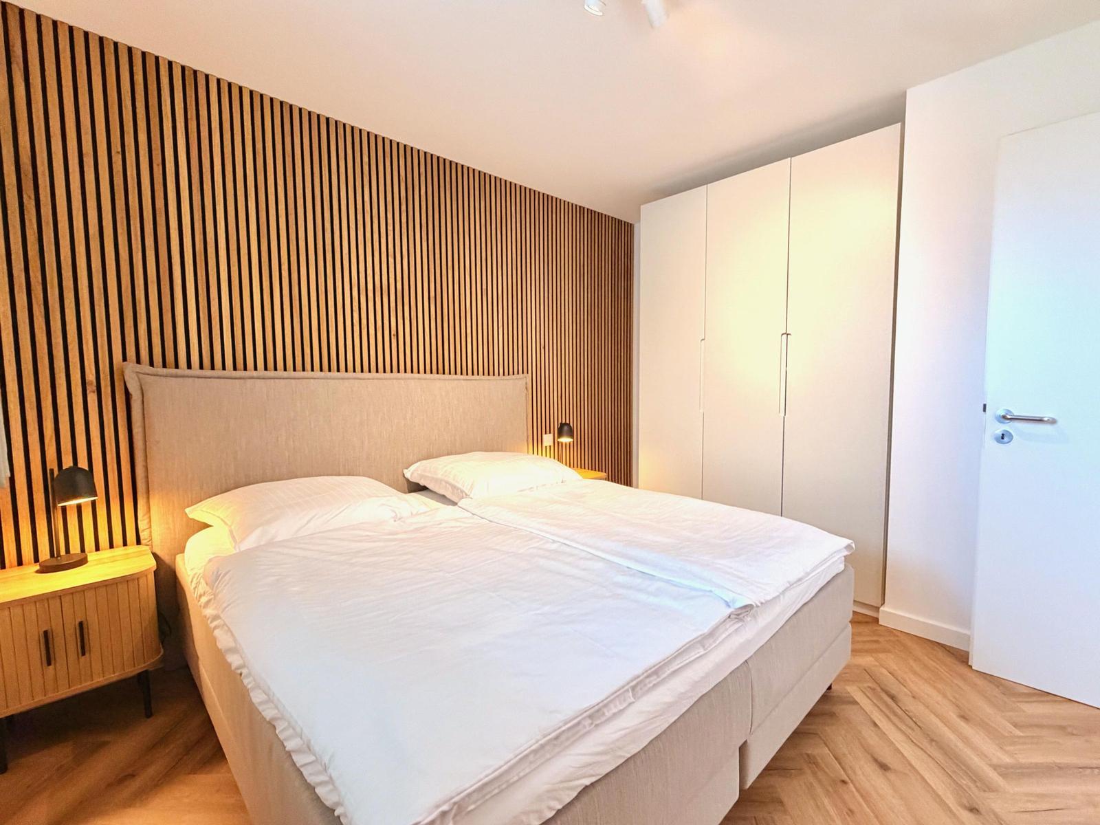 Schlafzimmer mit Doppelbett, Holzwand und Schrank. Holzdielenboden und Nachttische mit Lampen.
