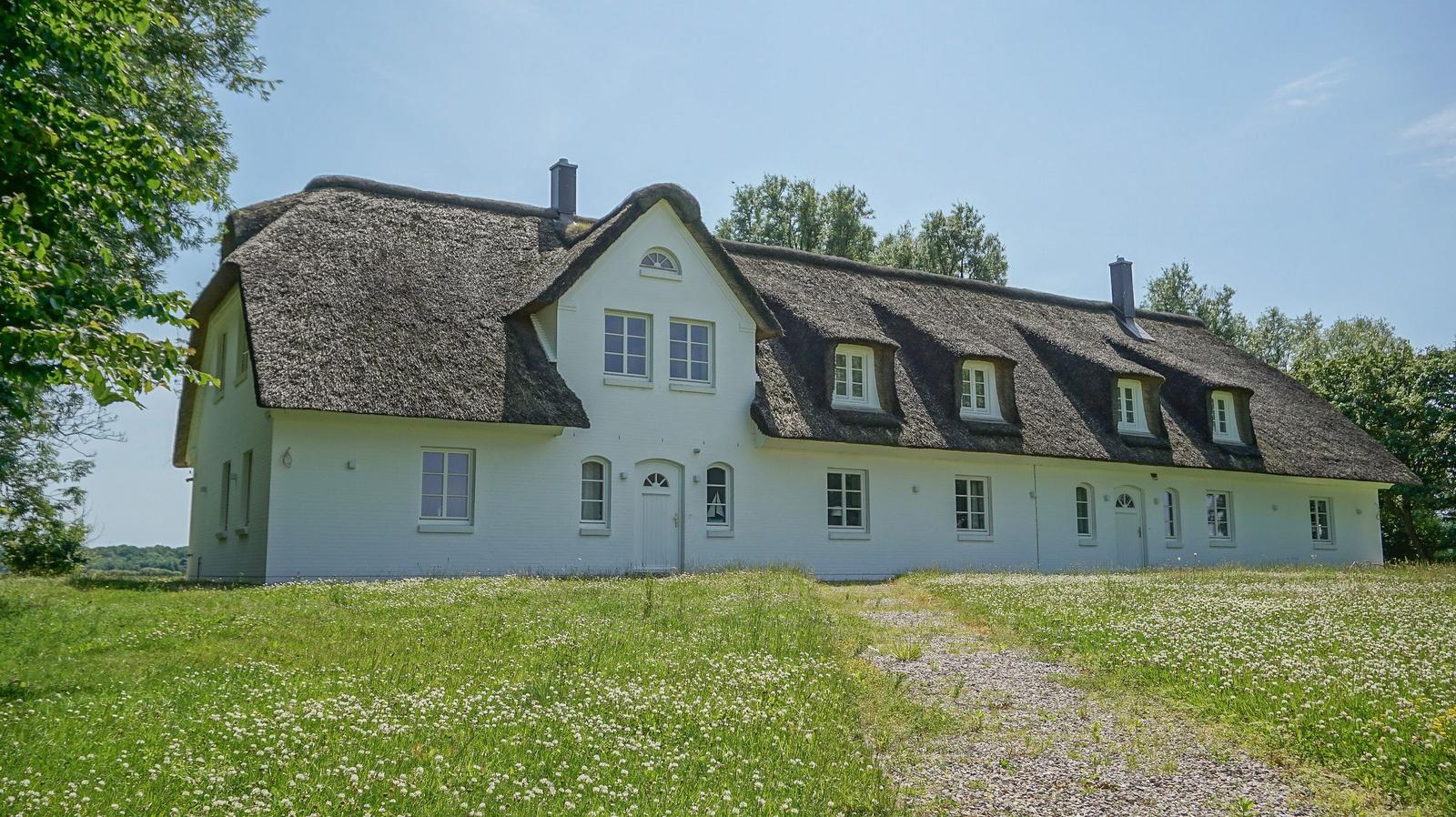 Weißes Haus mit strohgedecktem Dach und mehreren Fenstern in grüner Umgebung.