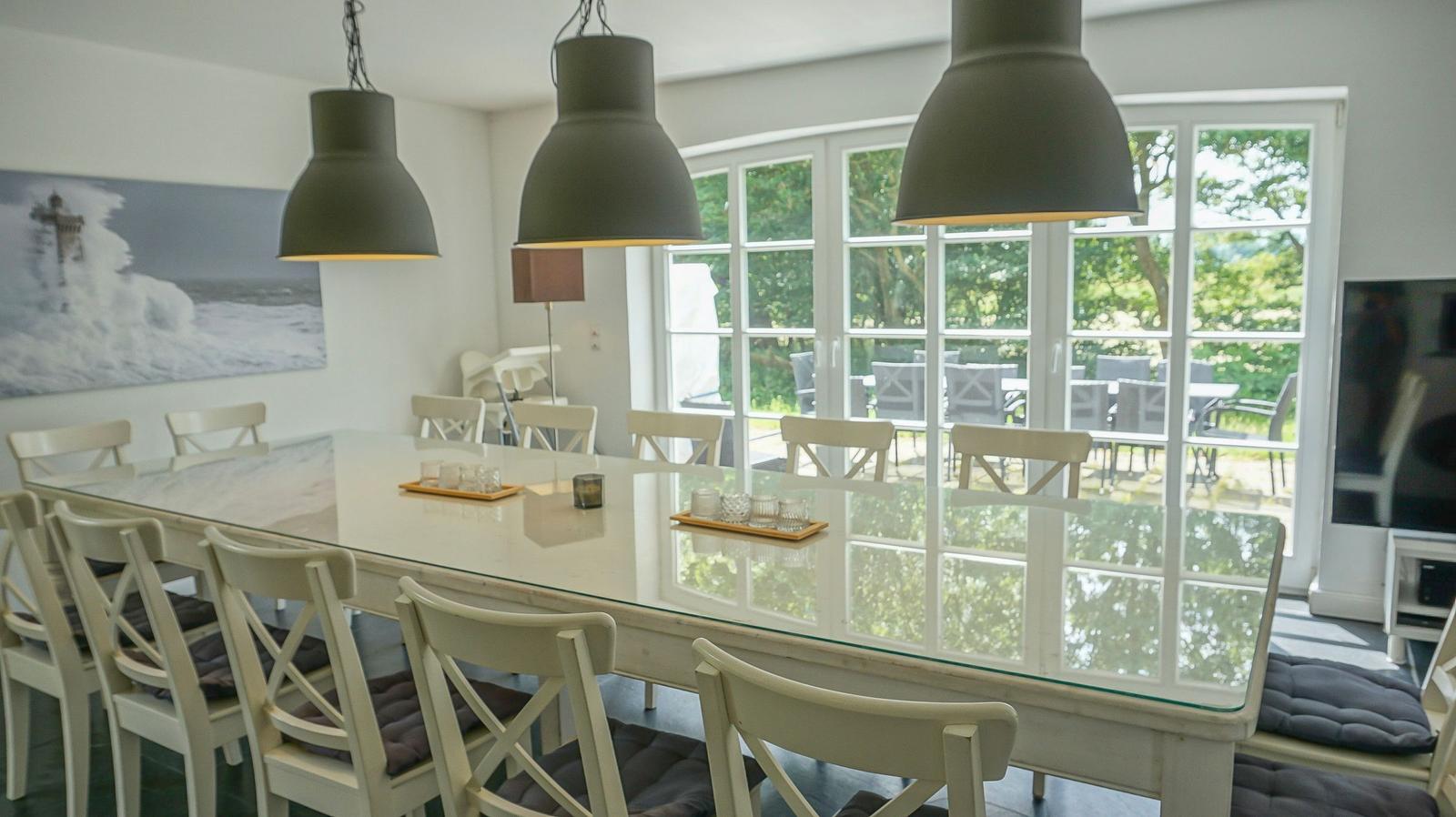 Esszimmer mit langem Tisch, weißen Stühlen und großen Fenstern zur Terrasse.
