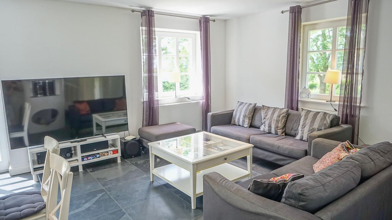 Wohnzimmer mit Sofa, Glas-Tisch und Fernseher. Fenster mit Vorhängen.