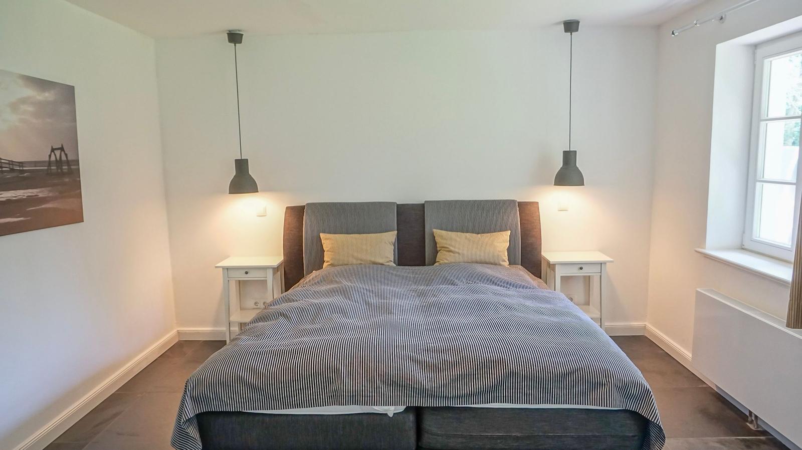 Schlafzimmer mit Bett, zwei Nachttischen und Hängelampen. Fenster und Wandbild sichtbar.