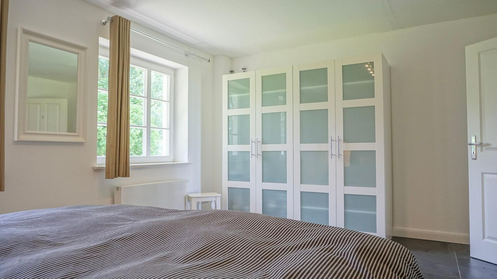 Schlafzimmer mit Bett, Kleiderschrank und Fenster mit Blick auf Grün.