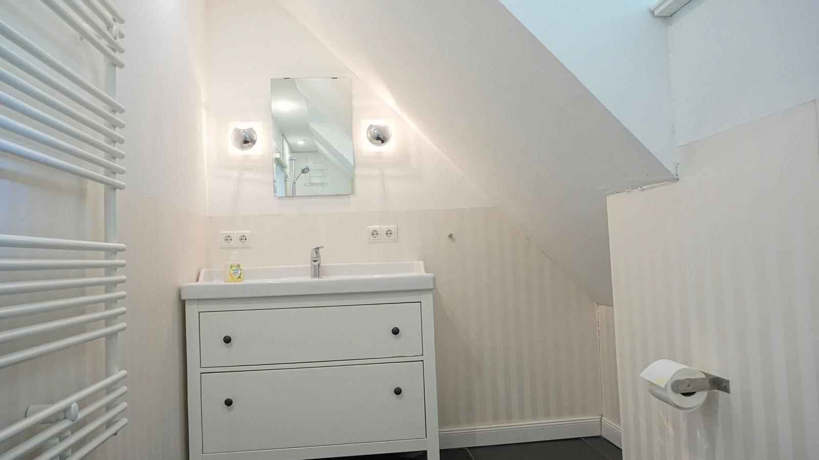 Badezimmer mit Waschbecken, Spiegel und Heizung unter schräger Decke.