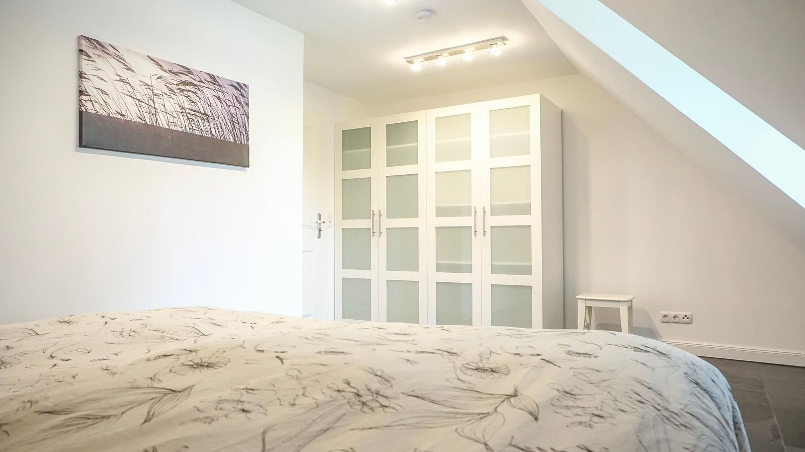 Schlafzimmer mit Dachschräge, weißer Kleiderschrank und Bett mit Musterdecke.
