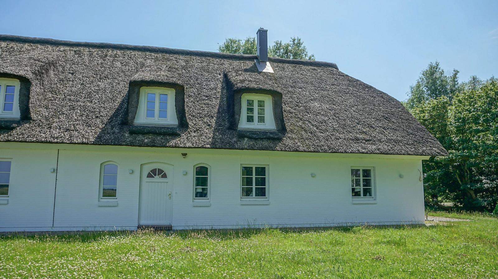 Ein weißes Haus mit Strohdach und mehreren Fenstern steht auf einer Wiese.