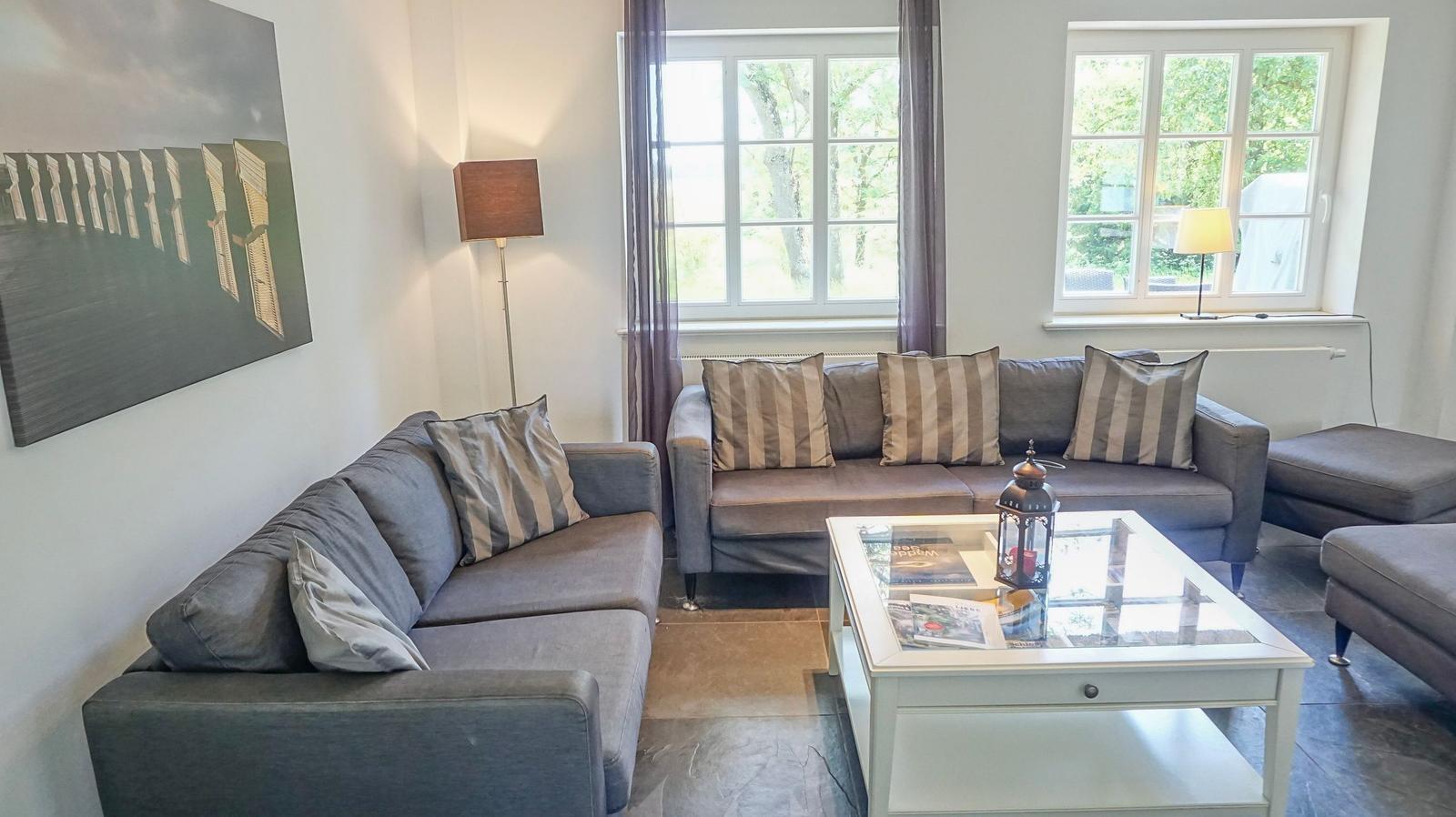 Wohnzimmer mit zwei grauen Sofas, Glas-Tisch und Fenstern mit Blick auf Grün.