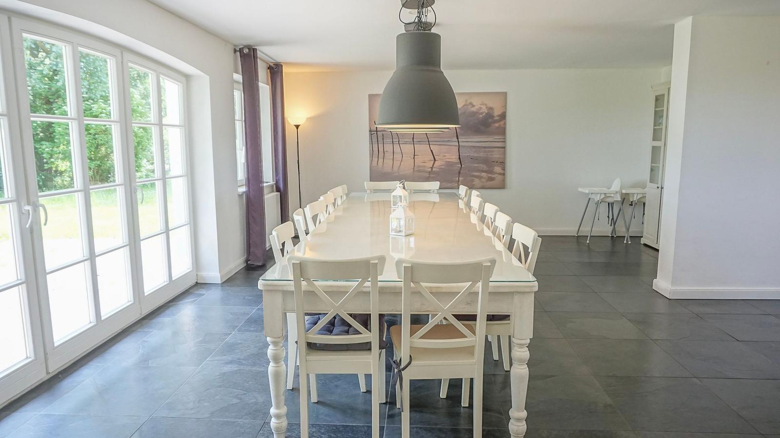 Esszimmer mit langem Tisch, weißen Stühlen und großem Fenster zur Außenseite.