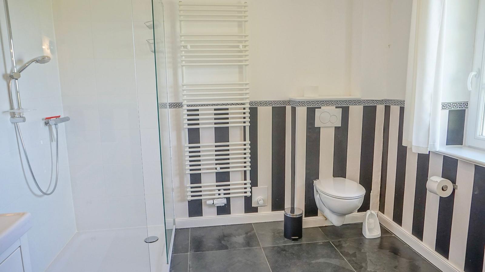 Bad mit Dusche, Toilette und Heizkörper an schwarz-weiß gestreifter Wand.