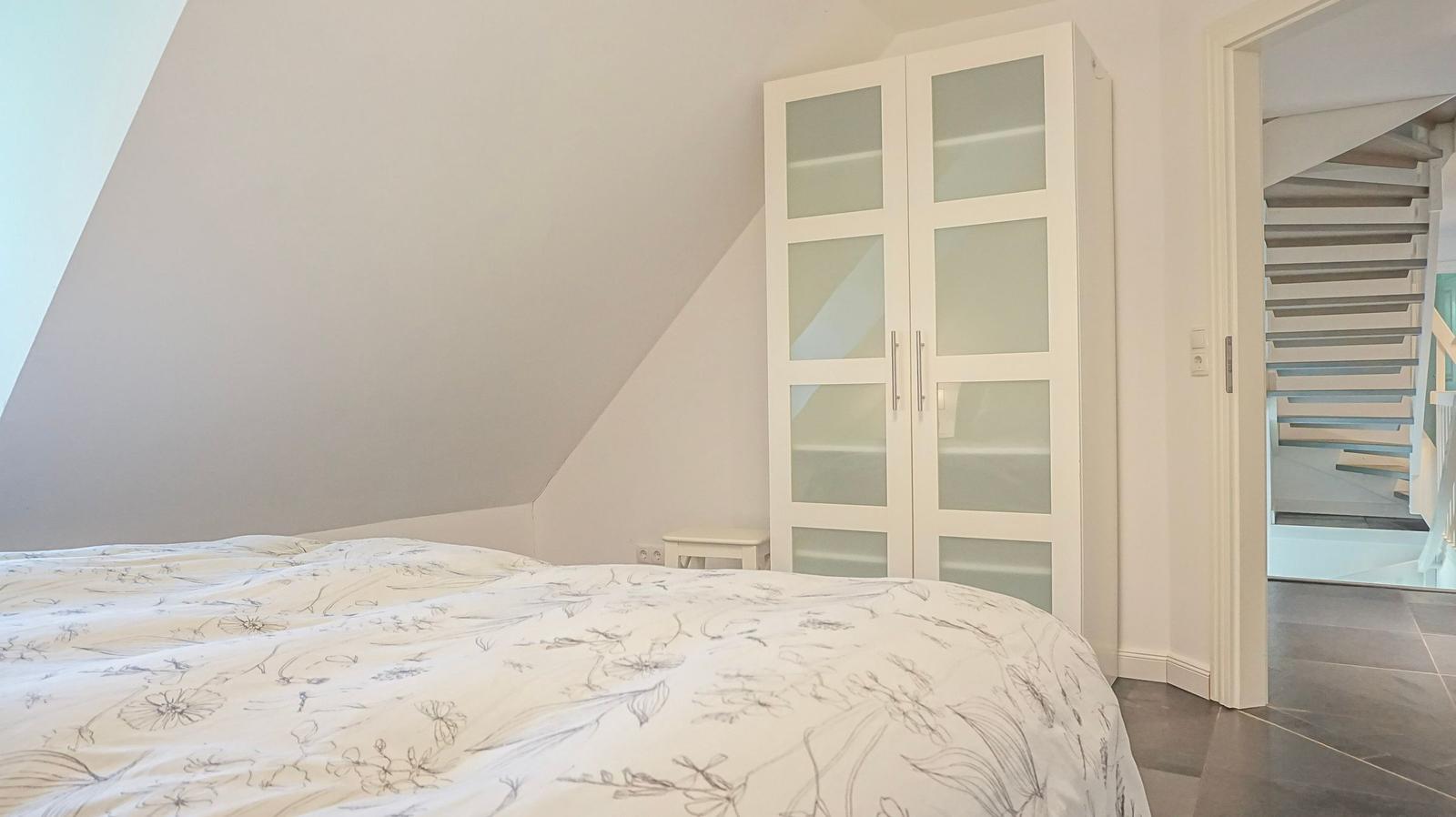 Schlafzimmer mit Dachschrägen, weiße Kommode und Blick auf Treppe.