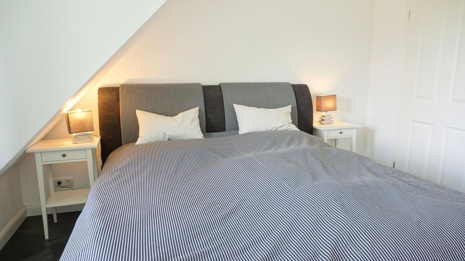 Schlafzimmer mit Bett, zwei Nachttischen und Lampen unter schräger Decke.