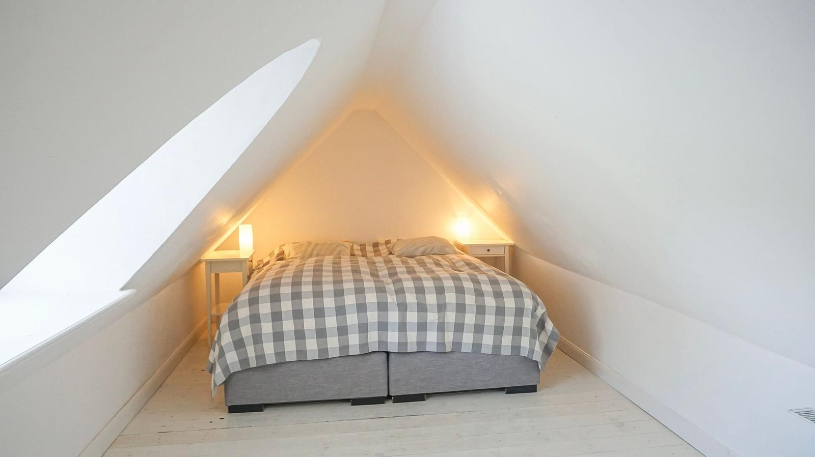 Schlafzimmer mit Dachschrägen, Bett mit kariertem Bettbezug und zwei Nachttischen mit Lampen.