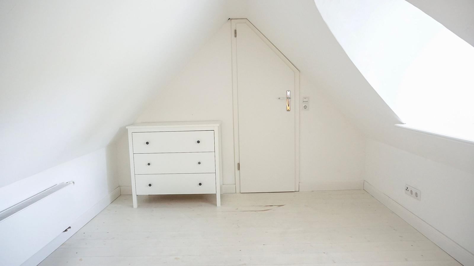 Zimmer mit Dachschrägen, weißer Kommode und Tür.