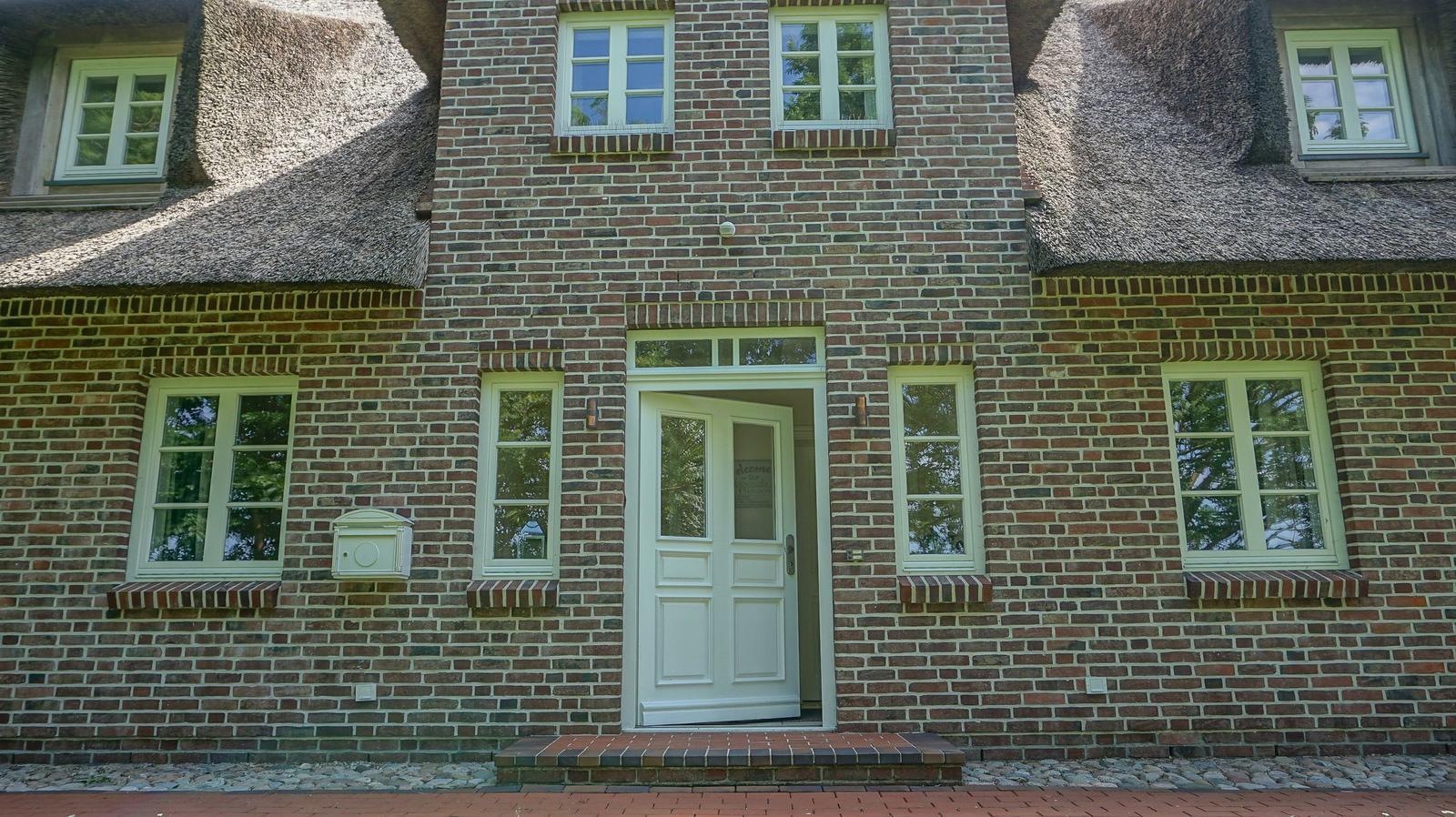 Haus mit Backsteinfassade, Strohdach und weißen Fenstern. Eingangstür mit Glasfenstern.