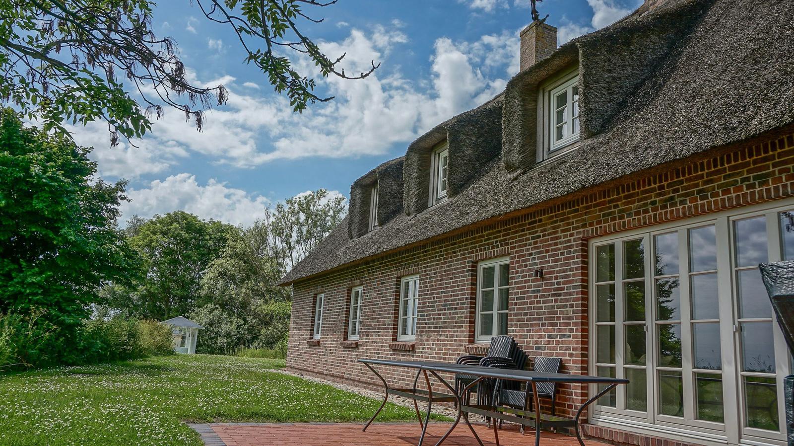 Haus mit Strohdach, Terrasse und Garten unter blauem Himmel.