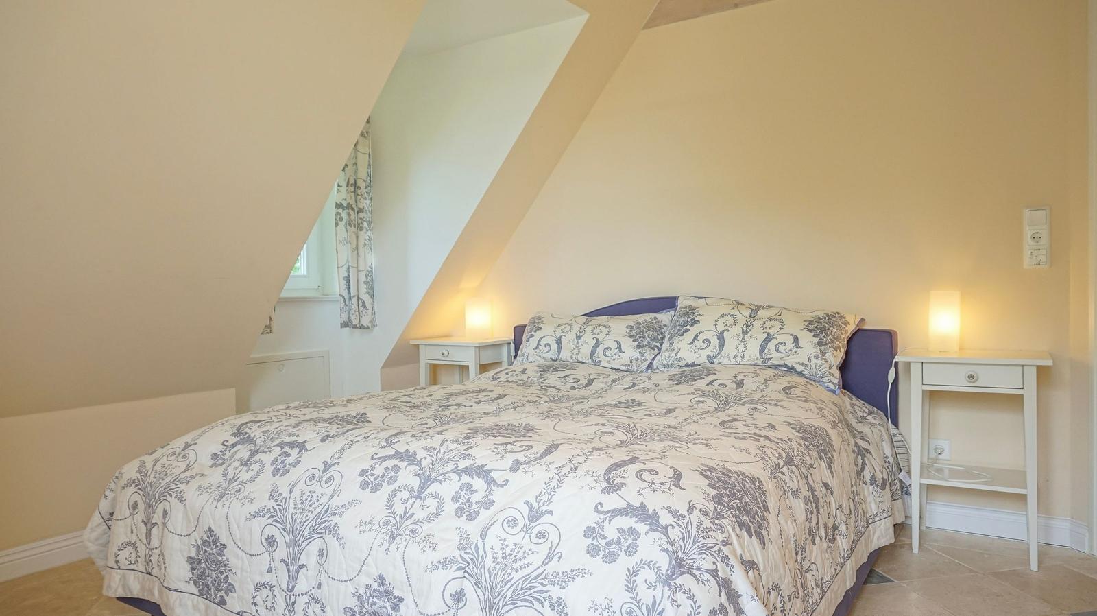 Schlafzimmer mit Dachschrägen, Bett mit gemustertem Bettbezug und zwei Nachttischen mit Lampen.