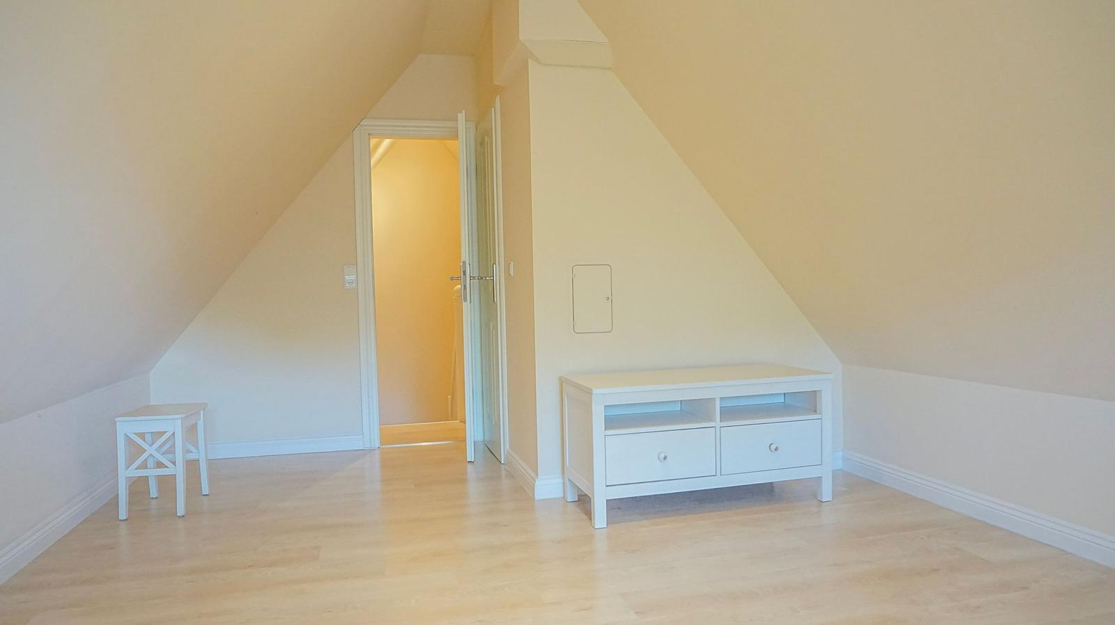 Zimmer mit schrägen Wänden, weißem Schrank und Holzboden. Tür führt in einen anderen Raum.