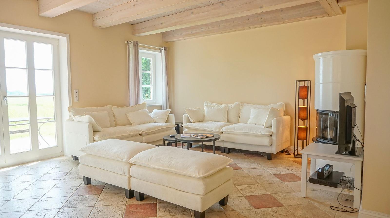 Wohnzimmer mit weißen Sofas, Holzbalken und Terrassentür. Kamin und TV-Tisch sichtbar.