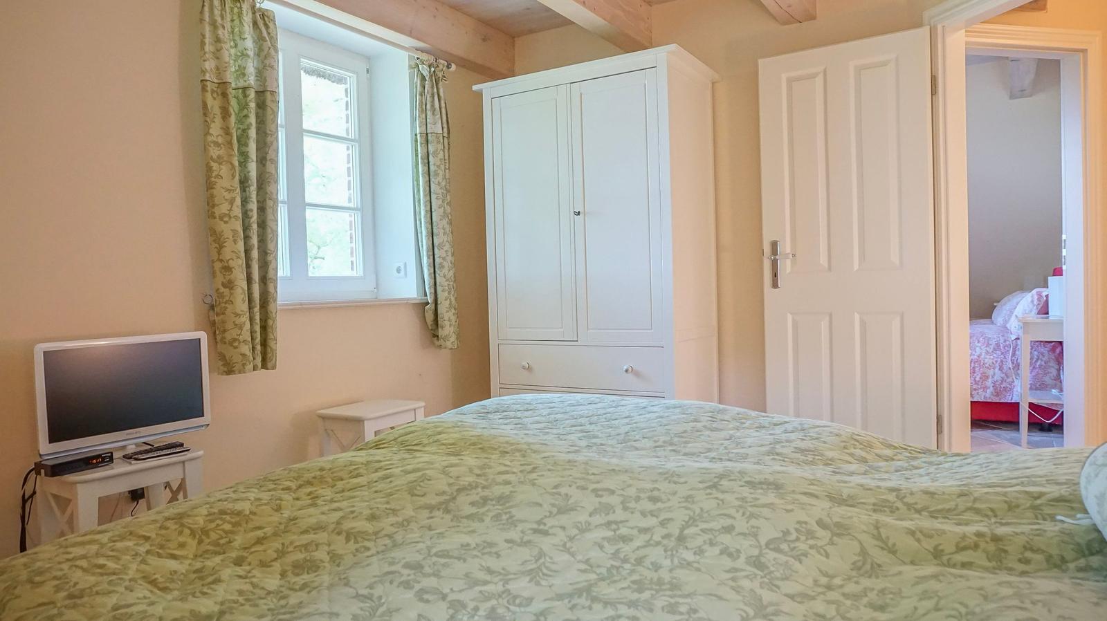 Schlafzimmer mit Bett, Kleiderschrank und Fenster. Tür führt in ein weiteres Zimmer.
