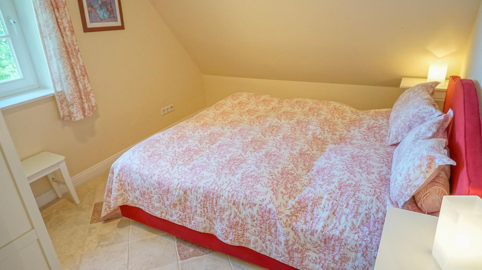 Schlafzimmer mit Dachschräge, Bett mit rosa-weißem Muster und Fenster mit Vorhängen.
