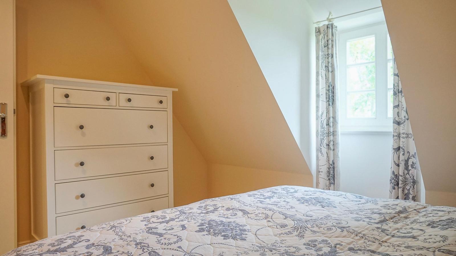Schlafzimmer mit Dachschrägen, weißer Kommode und Bett mit floraler Bettwäsche.
