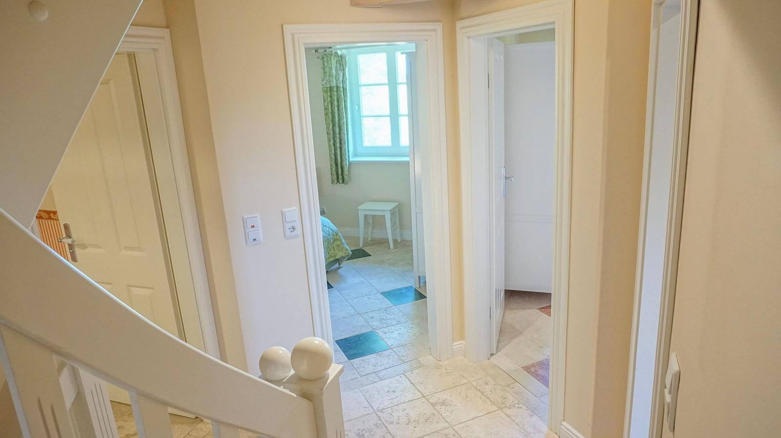 Hausflur mit Treppe, Tür zu Schlafzimmer und Bad. Fliesenboden und Fenster mit Vorhang.