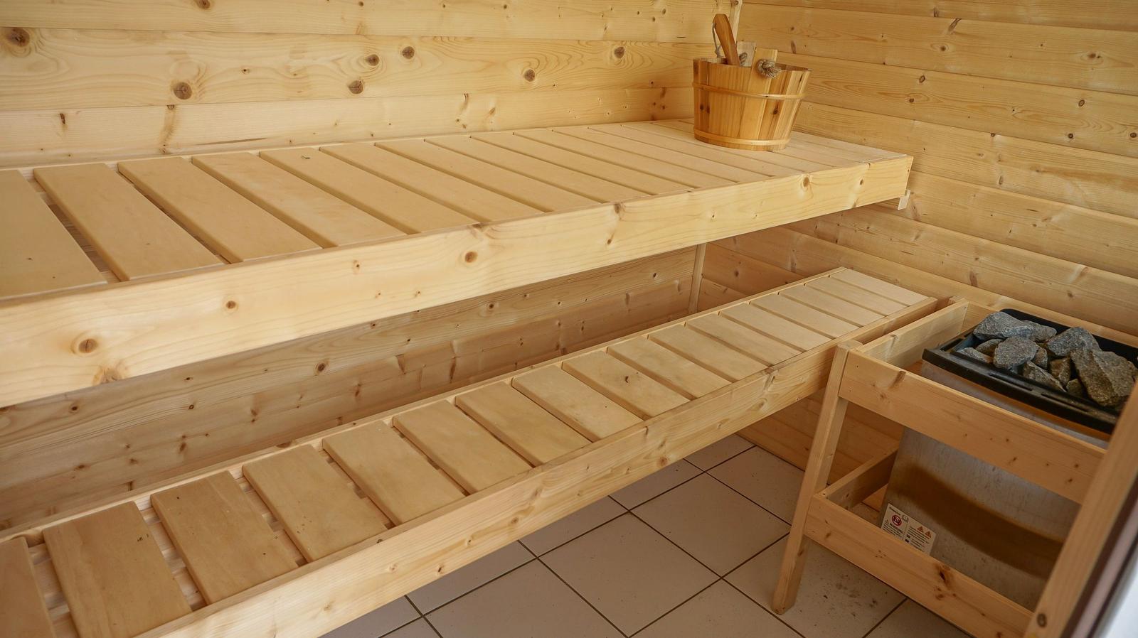 Sauna mit Holzbank und Steinofen. Holzgehäuse und Körbe sind sichtbar.