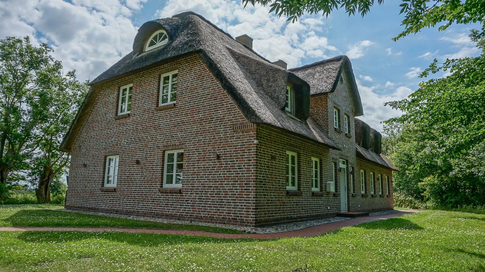 Rotes Backsteingebäude mit strohgedecktem Dach und weißen Fenstern in grüner Umgebung.