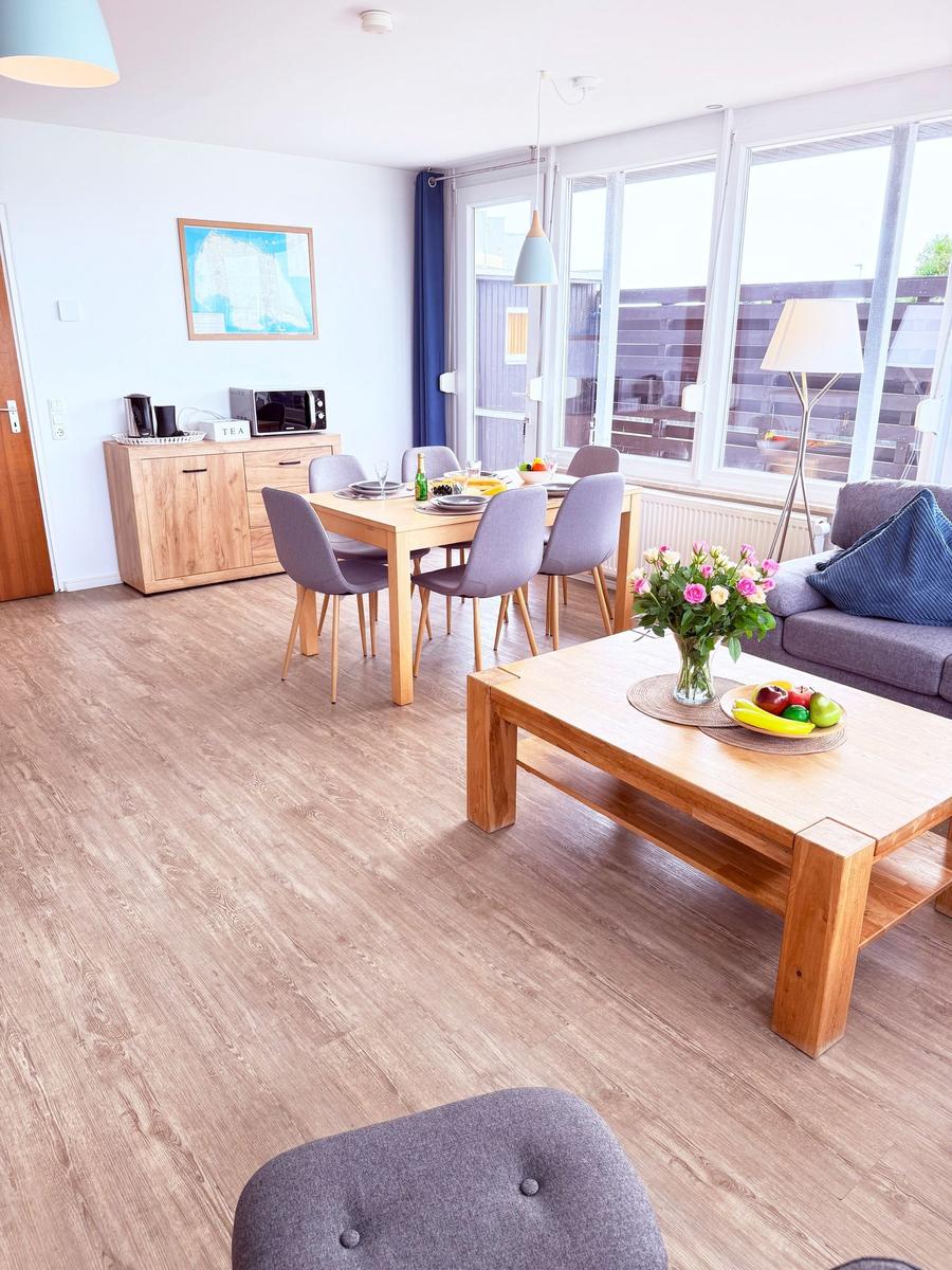 Wohn- und Essbereich mit Holzboden, Tisch, Stühlen und Sofa. Große Fensterfront mit Blick auf Balkon.